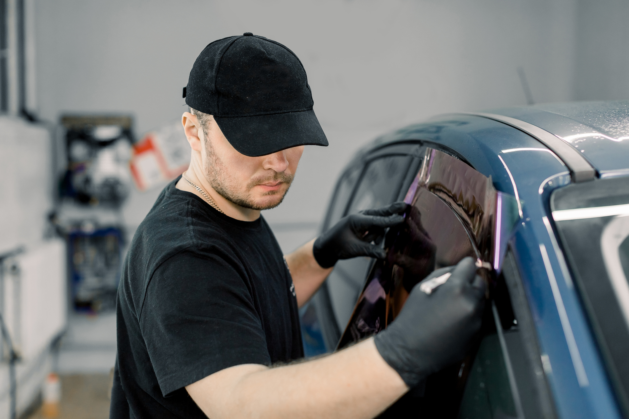Closeup image of a handsome car mechanic worker, wearing black uniform, attaching tinting foil to car window in specialized service station.